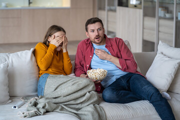Frightened spouses watching horror film series, sitting on couch. Emotional scared woman covering...