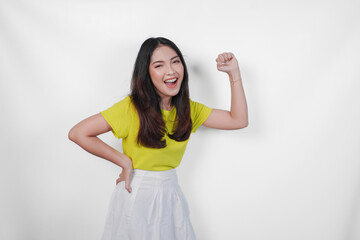 Confident Asian woman in a yellow shirt and white skirt is showing strong gesture by lifting her arms and muscles smiling proudly, isolated by white background.