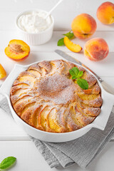 Delicious peach pie in a white ceramic baking form on a white wooden background. Summer cake. Selective focus, copy space.