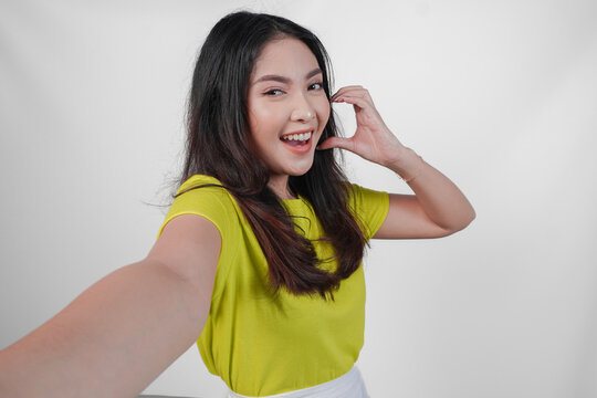 Cute young Asian woman in a yellow shirt is taking a selfie while making a heart gesture on her cheek, isolated over white background.