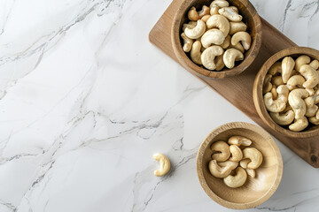 Fototapeta premium Top view of cashew nuts in wooden bowls on a kitchen board over a white marble background