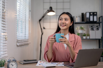 Young woman enjoying coffee at her desk, working from home with a laptop and documents, looking content and productive in a cozy home office setup.
