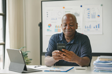 A man using his smartphone while working in a modern office with charts on the whiteboard and a laptop on the desk.