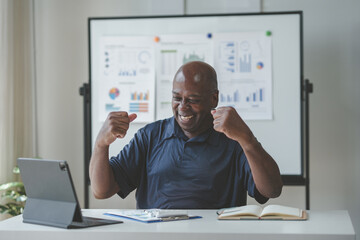 Excited businessman celebrating success in front of a tablet and charts, demonstrating motivation and achievement in a modern office environment.