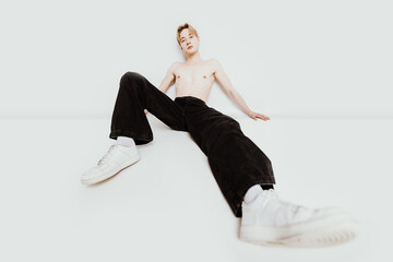shirtless handsome male model sits on the floor, looking at the camera with extremely long legs exaggerated by the camera angle, striking a high fashion, modern art pose