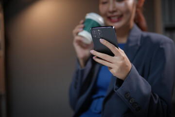 Businesswoman smiling, holding coffee and using smartphone in modern office setting. Relaxed, professional attire and environment.