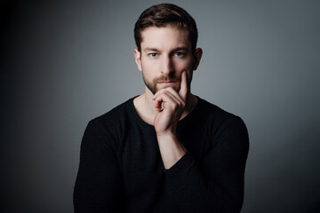 Handsome Young Man in Black Sweater Posing Thoughtfully with Hand on Chin Against Gray Background while finger is high pointing to the top and face straight to the camera