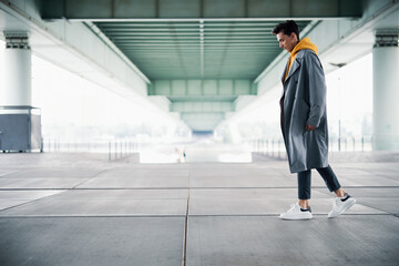 Modern Handsome Man in Gray Coat and Yellow Hoodie Walking Under a Bridge, Looking Down Thoughtfully