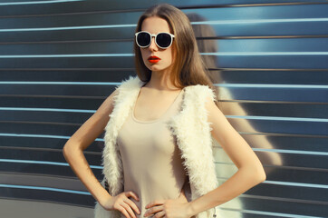 Modern stylish young woman in white fur vest, glasses, fashionable girl posing on gray background