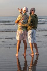 gay couple back to back with their dog on their lap looking at the camera, on the beach