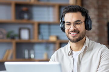 Fototapeta premium Smiling young Muslim man working and studying remotely at home. He sits in headphones at the laptop and smiles at the screen