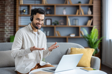 Smiling young Indian man sitting on sofa at home wearing headset, talking on video call, gesturing with hands