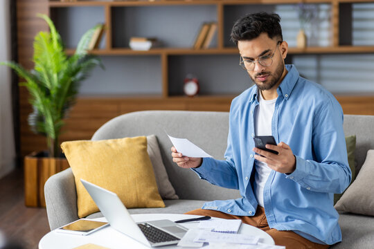 Worried young Indian man working from home on laptop, seriously checking and holding documents and mobile phone