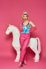A woman in pink pants, a sequined top, and a crown poses in front of a white unicorn.