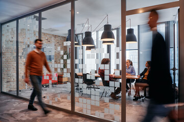 Fototapeta premium In the dynamic hustle and bustle of a business environment, a group of young business professionals walk down the corridor next to their office, while their colleagues collaborate inside, symbolizing