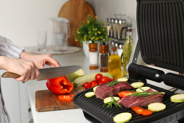 Woman cooking different products with electric grill at white wooden table in kitchen, closeup
