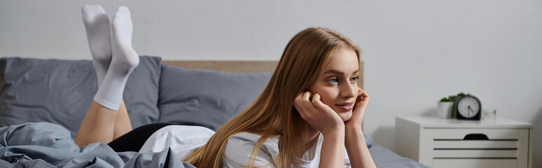 A young woman lays in bed, lost in thought, with her feet up in the air.
