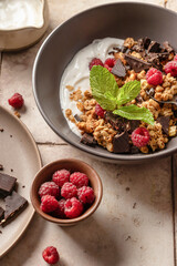 Granola with yogurt, chocolate, fresh raspberries and mint on beige tile background. Organic food for healthy and dieting breakfast