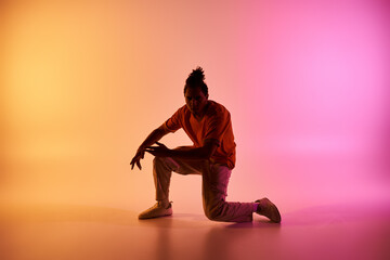 A young African American man in a vibrant orange shirt dances in a studio, illuminated by a...