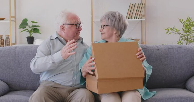 Elderly couple is delighted with surprise when they unpack package together in cozy home. Mature couple is sitting on sofa and opening cardboard box with excited and joyful expressions on their faces.