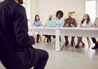 Fototapeta premium Coach and motivated employees sitting at desk listening at training, presenting workshop, engaged audience. Business leader interacting team at corporate meeting in office to study, people discussing