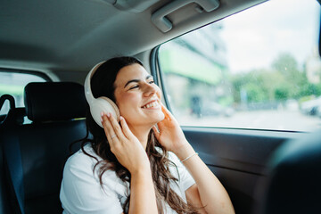 One young beautiful caucasian woman is listening to music on wireless headphones on backseat in the car