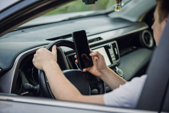 Casual driver guy distracted by his phone while in front of the steering wheel, using his smartphone with one hand while driving. Risk and danger situations on the road, violating traffic rules