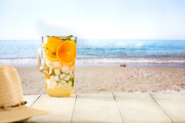 Glass of mocktail drink with ice and citrus fruit on a wooden table with sandy tropical beach and blue sky background. Summer table and copy space for products and food and drinks and fruit.