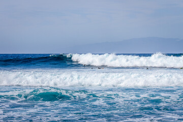 Waves coming ashore on a sunny day and surfers ready to surf the waves. Wall of blue wave with white foam coming ashore where surfers are surfing. Atlantic Ocean. Tenerife, Canary Islands, Spain.