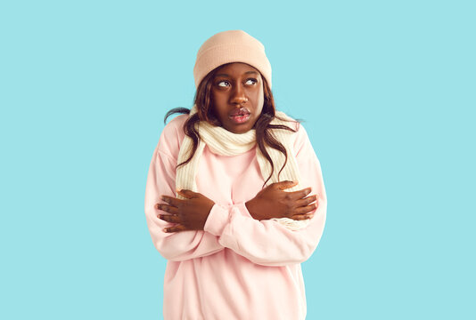 Portrait of unhappy African American woman who is freezing because of cold weather and can't keep warm. Dark-skinned woman in sweatshirt, scarf and hat is shivering from te cold on pink background.