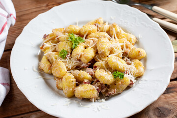 Gnocchi (small potato balls) with bacon in creamy sauce in a white plate . Close up view. Lunch table background, fork, garlic. Traditional Italian cuisine.