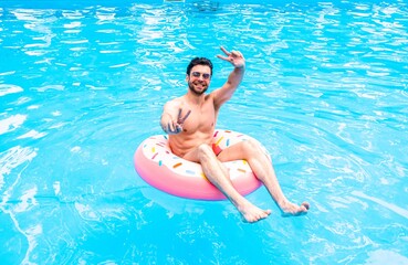 Happy guy in sunglasses showing victory sign and resting in the swimming pool with inflatable ring outdoors. Summertime, holidays, lifestyle concept