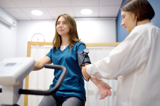 Female doctor cardiologist measures pressure with patient during bicycle exercise for examination cardiovascular system at medical office of clinic. Young woman training on bike simulator. - Powered by Adobe