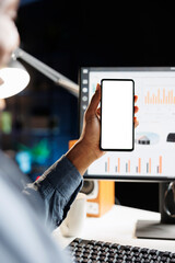 Remote employee uses phone with white screen at workstation, examining business sales target on online statistics. Young man holds mobile device running isolated blank display at home.