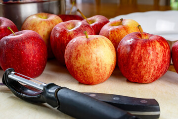 A knife and peeler on a white cutting board surrounded by gala apples.  Preparation for a pie..
