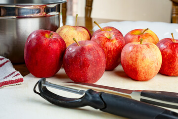 A knife and peeler on a white cutting board surrounded by gala apples.  Preparation for a pie..
