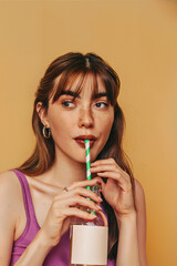A young woman with brown hair and freckles, sipping a beverage through a green and white striped straw from a clear bottle. Blank white label mockup.