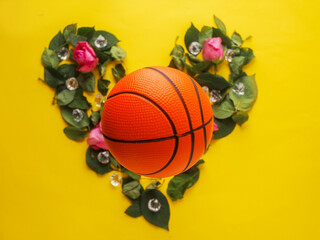 Classic Basketball on green leaf in heart shape and red roses and gem stones, yellow color surface background. Love sport concept. Flat lay.