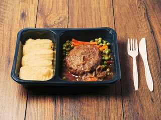 A meal consisting of burger, mashed potatoes and vegetables in plastic tray is served on wooden table and plastic fork and knife. Simple classic dish for take away. Office lunch.