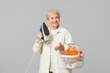 Happy senior woman with iron and basket of laundry on grey background