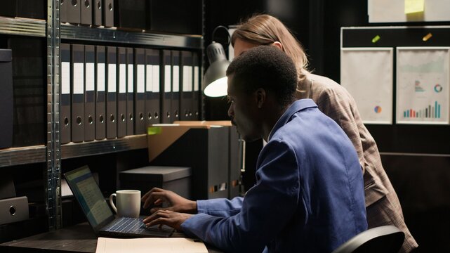 Caucasian and african american law enforcement personnel and investigators meticulously analyze evidence and exchange information in office. Black policeman and his female police partner review cases.