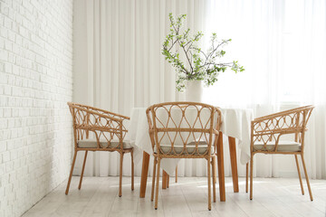 Vase with blooming branches on table near armchairs in white dining room
