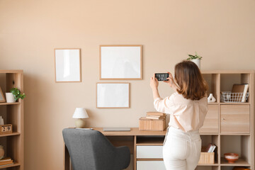 Young woman taking picture of hanging blank frames on beige wall at home