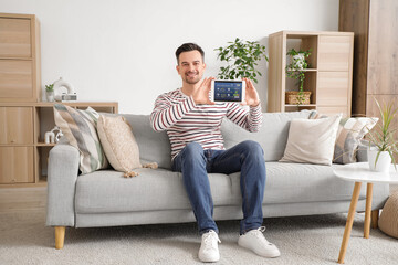 Young man with smart home security system control panel sitting on sofa in room