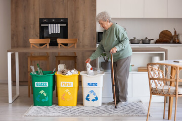 Senior woman throwing plastic bottle into garbage bin in kitchen. Waste sorting concept