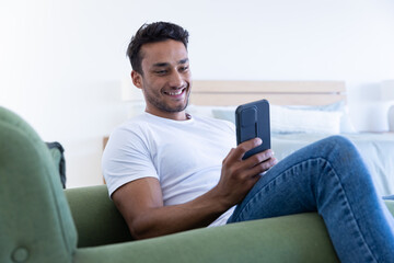 Smiling man relaxing on green sofa, using smartphone for entertainment at home