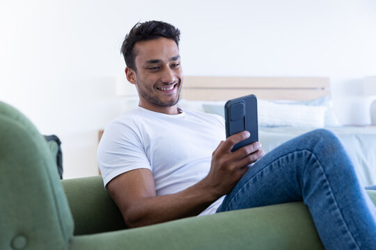 Smiling man relaxing on green sofa, using smartphone for entertainment at home