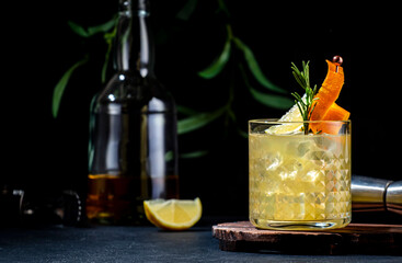 Yellow alcoholic cocktail with whiskey, honey ginger, lemon, juice, rosemary and ice in glass garnished with orange zest. Black background