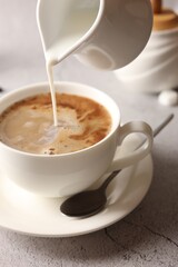 Pouring milk into cup with coffee on light grey textured table, closeup