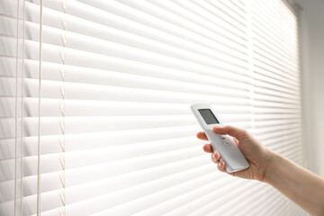 Woman using remote control to adjust window blinds indoors, closeup. Space for text
